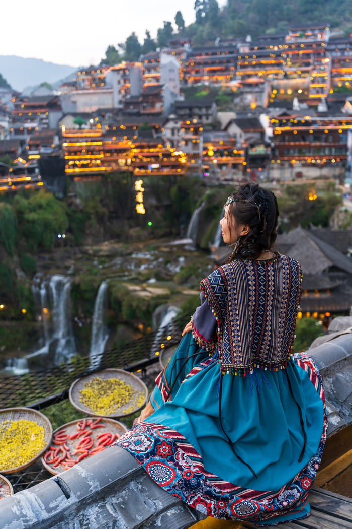 Mujer tradicional china de espaldas, sentada sobre el borde de un templo observando la ciudad montañosa, la cascana y vendiendo comida en Li Jiang, China.